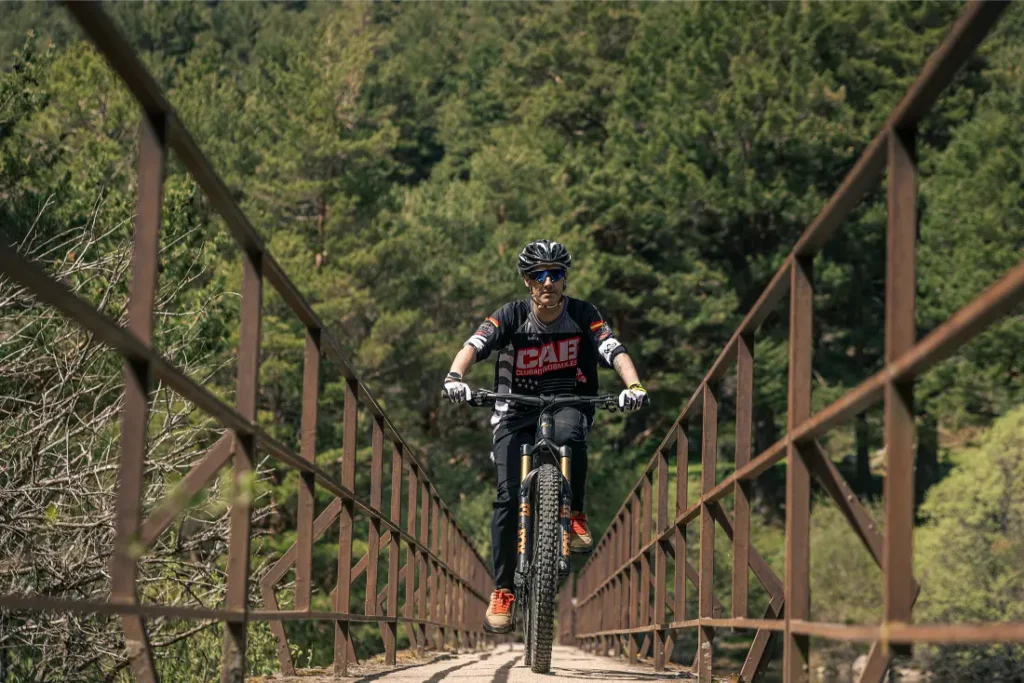 Una persona con casco y gafas de sol conduce una bicicleta de montaña con más potencia, cruzando un estrecho puente metálico rodeado de densos árboles verdes.