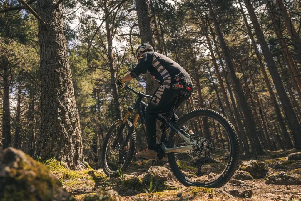 Una persona con casco conduce una bicicleta de montaña cuesta arriba por un sendero rocoso a través de un denso bosque, aprovechando el control y la potencia de una Bosch Performance Upgrade mientras la luz del sol se filtra entre los árboles.