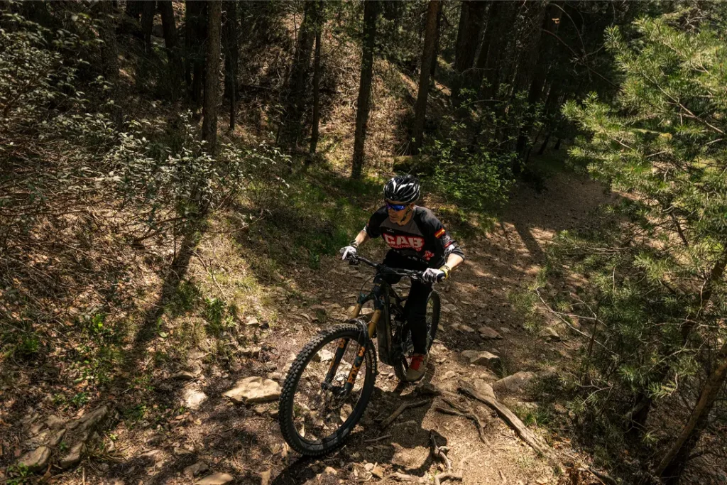 Un ciclista con casco conduce una bicicleta de montaña cuesta arriba por una pista forestal rocosa, disfrutando de más potencia y una experiencia sobre la bici mejorada gracias al Bosch Performance Upgrade 2.0, rodeado de árboles y la luz del sol.