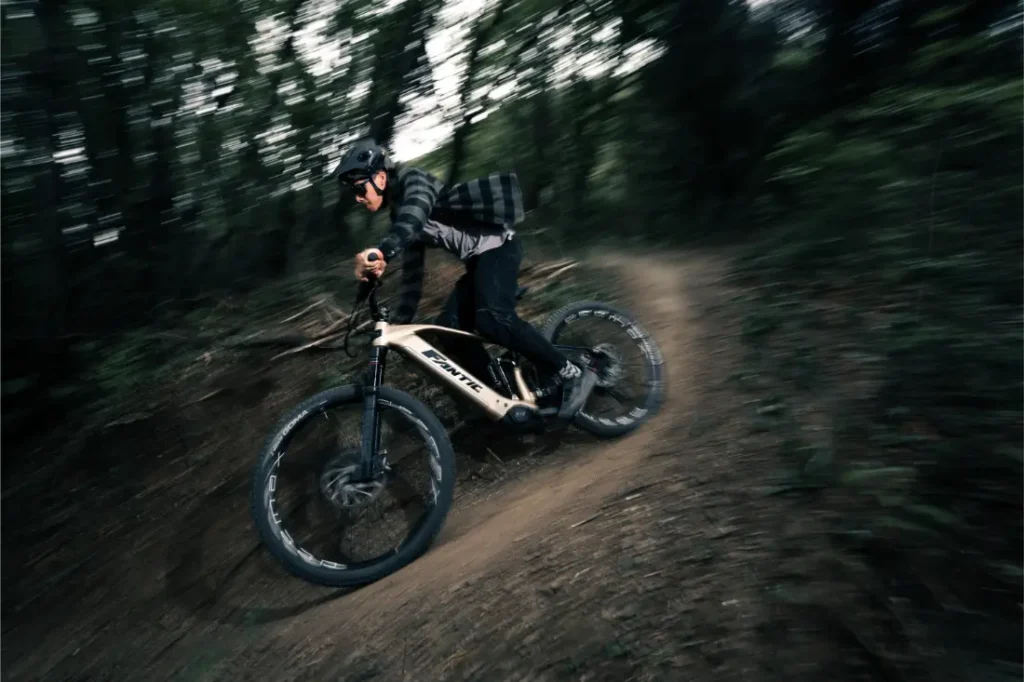 Una persona con casco conduce rápidamente una Fantic Sierra e-MTB por un sendero de tierra en una zona boscosa, rodeada de árboles y vegetación.