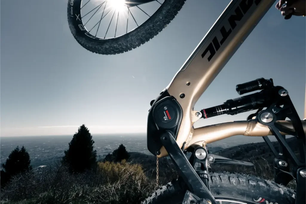 Primer plano del cuadro de una Fantic Sierra e-MTB y el motor Bosch CX Gen5, con la rueda trasera levantada y la luz del sol brillando a través de la rueda sobre un pintoresco telón de fondo al aire libre.