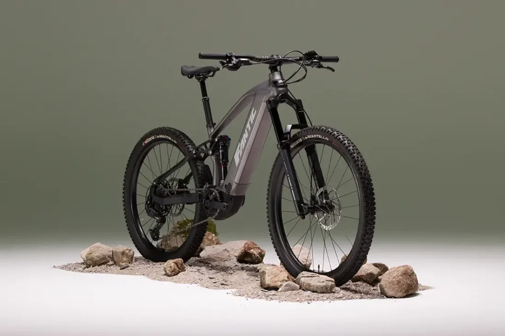Una Fantic Sierra e-MTB gris mate, propulsada por el motor Bosch CX Gen5, se muestra sobre grava y rocas sobre un fondo verde apagado.