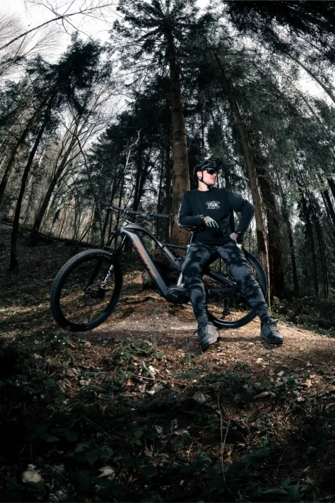 Persona con ropa de ciclismo y casco negros junto a una Fantic Sierra e-MTB, con motor Bosch CX Gen5, en una zona boscosa con árboles altos y el suelo cubierto de hojas.