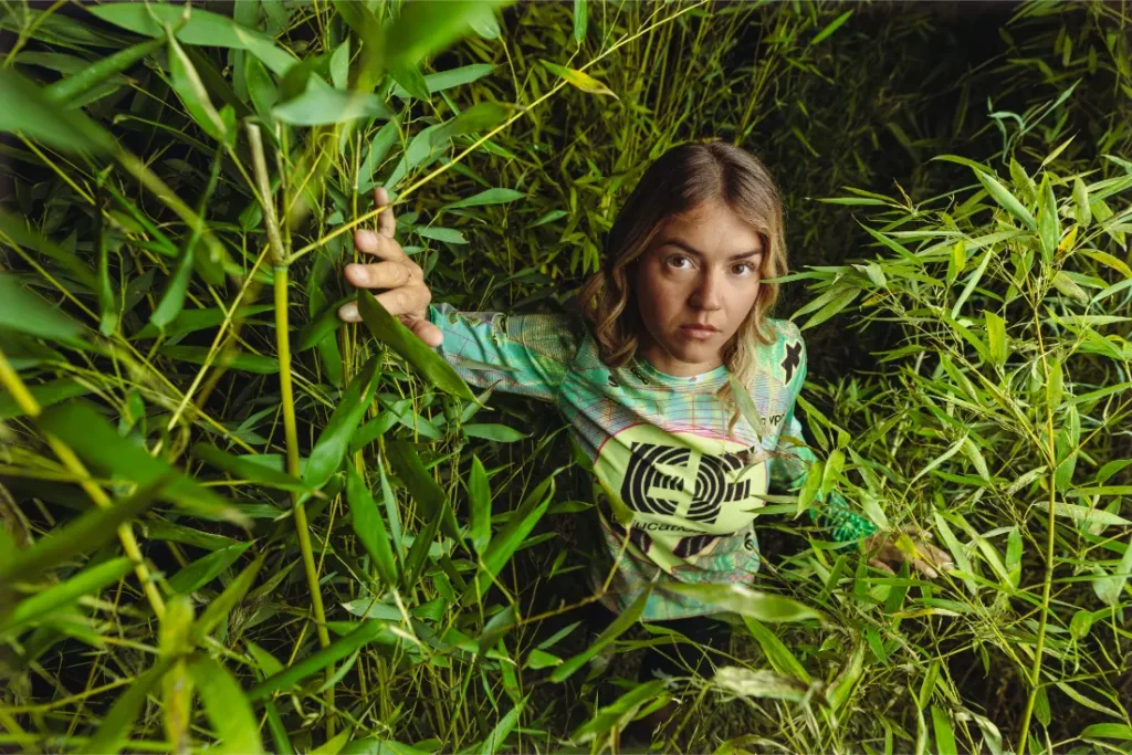 Una persona se encuentra entre densas plantas verdes de bambú, agarrada a una rama y mirando hacia la cámara con expresión neutra, luciendo un maillot ASSOS del equipo EF Pro Cycling, un sutil guiño a las aspiraciones del Giro 2026.