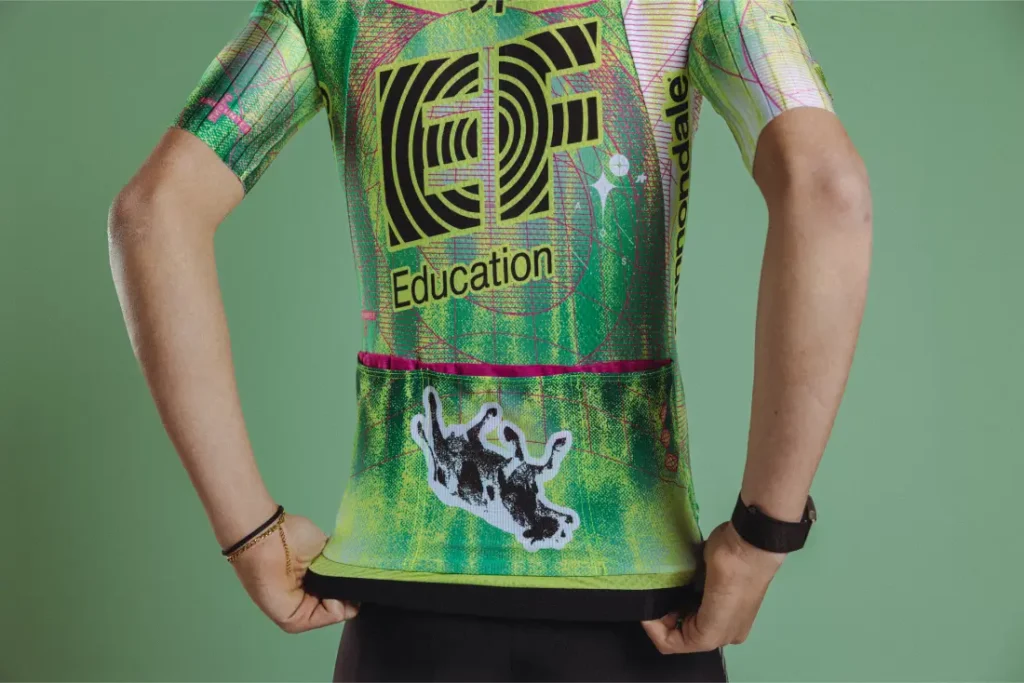 Persona que lleva un maillot verde EF Pro Cycling de ASSOS, con un gráfico de una vaca cayendo en la espalda, sobre un fondo verde liso.
