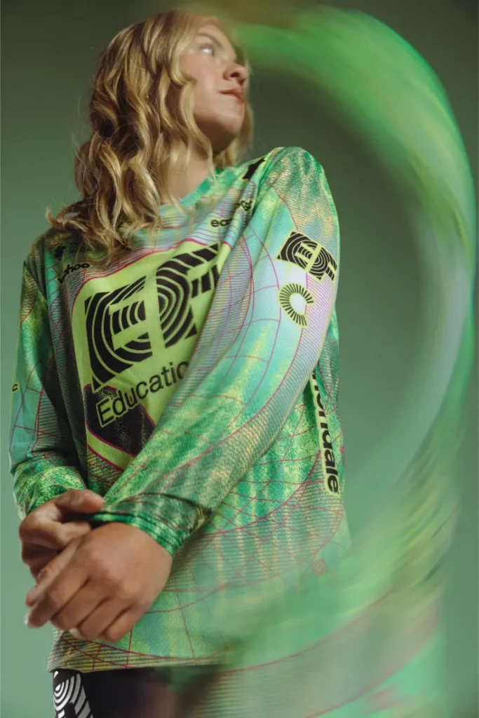 Una persona vestida con una camiseta ASSOS verde de manga larga con llamativos gráficos negros mira hacia un lado sobre un fondo verde. Un remolino verde aparece en primer plano, aludiendo a la energía dinámica de EF Pro Cycling para el Giro 2026.