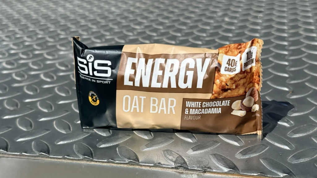 SiS Energy Oat Bar, una barrita energética de chocolate blanco & macadamia con 40g de carbohidratos, reposa sobre una superficie metálica texturizada bajo la luz del sol.