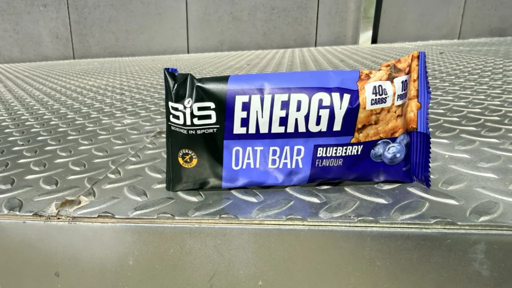 A barrita energética SiS de avena sabor arándano descansa sobre una superficie metálica, mostrando en su etiqueta el aporte de 40g de carbohidratos resaltado en el envase.