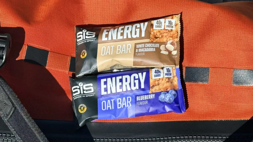 Dos barritas energéticas SiS Energy Oat Bars, una de chocolate blanco y macadamia y otra con sabor a arándanos, se asientan sobre una superficie naranja con una correa negra y una tira reflectante, ideal para un sabroso chute de carbohidratos.