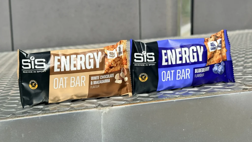 Two SiS Barritas energéticas de avena, una de proteína sabor chocolate blanco y macadamia y otra de arándanos, se exhiben sobre una superficie metálica.
