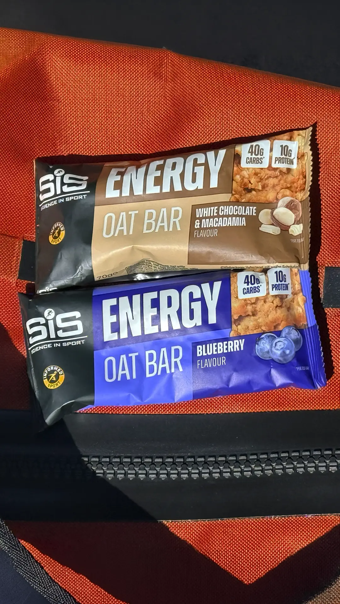Two SiS barritas energéticas de avena, una de chocolate blanco y macadamia y otra de arándanos, ricas en carbohidratos, se encuentran sobre una tela naranja.