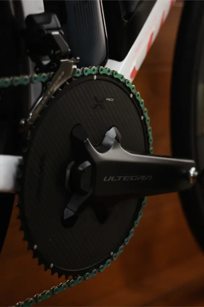 Primer plano de un plato de bicicleta con un brazo de biela Shimano Ultegra, un plato de fibra de carbono Aurora Green y una cadena KMC X12L Waxed para una cadena eficiente.