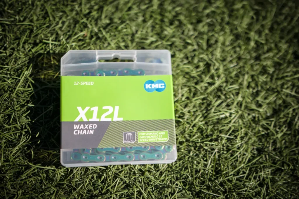 Una cadena de bicicleta KMC X12L Waxed de 12 velocidades en un embalaje de plástico transparente descansa sobre césped artificial verde, mostrando sus detalles en verde Aurora y su eficiencia silenciosa para una conducción suave y silenciosa.