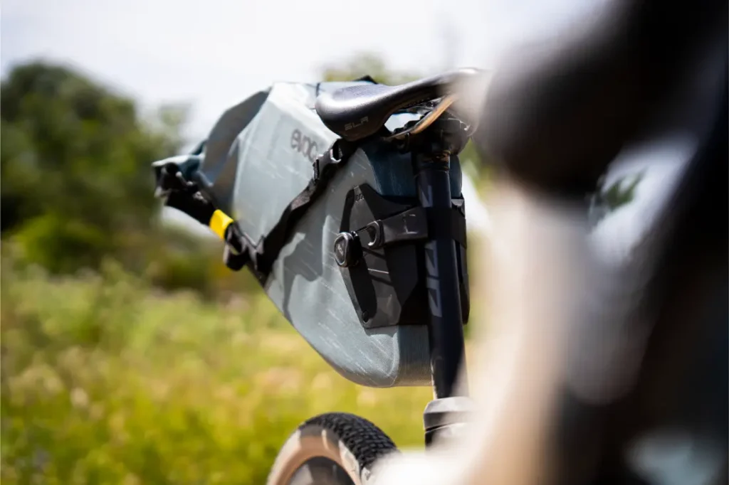 Primer plano de un sillín de bicicleta con una gran bolsa gris EVOC Bikepacking colocada debajo, que ofrece protección adicional, al aire libre con vegetación borrosa de fondo.