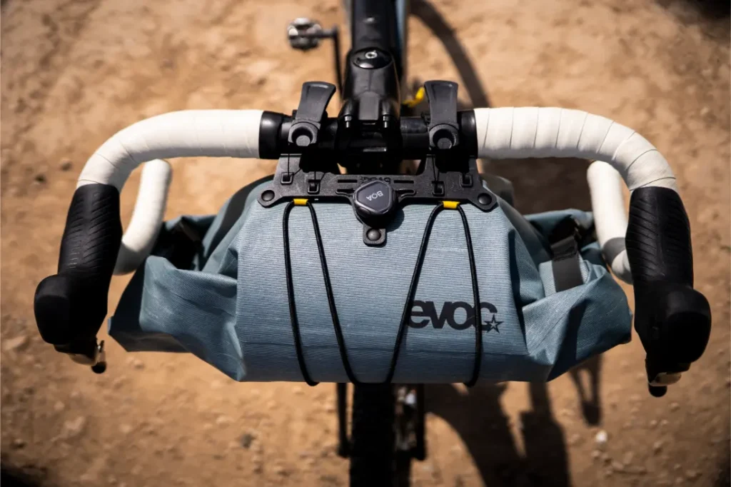 Primer plano del manillar de una bicicleta con una bolsa de manillar EVO gris acoplada, perfecta para aventuras en bicicleta, fotografiada al aire libre sobre una superficie de tierra.