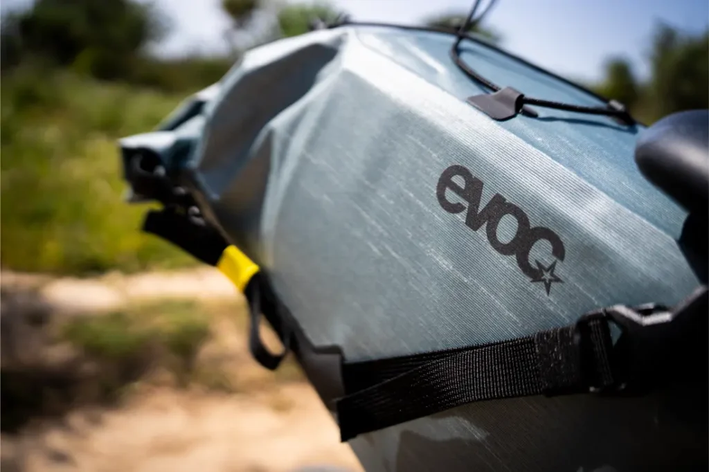 Primer plano de una bolsa para bicicleta de color gris azulado con el logotipo "evoc", sujeta a una bicicleta con correas negras: una pieza esencial del equipo EVOC Bikepacking, perfecta para transportar tu equipo protegido al aire libre en un entorno natural.
