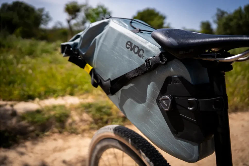Primer plano de un sillín de bicicleta con una gran bolsa gris EVOC Bikepacking sujeta a la tija del sillín, que ofrece protección adicional, colocado al aire libre en un día soleado con vegetación de fondo.
