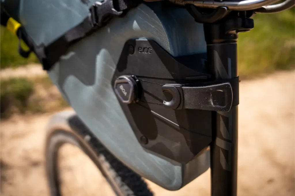 Primer plano de un cuadro de bicicleta con una bolsa de almacenamiento EVOC Bikepacking sujeta por un mecanismo de cierre negro cerca de la tija del sillín, que ofrece protección en sus paseos al aire libre por el camino de tierra.
