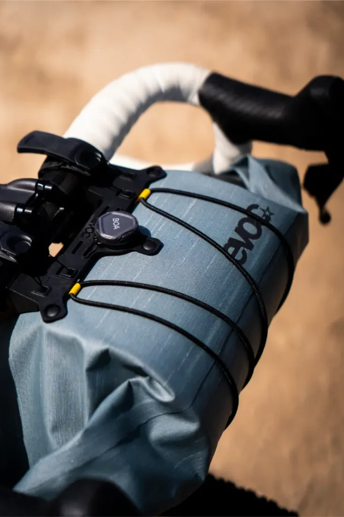 Primer plano de un manillar de bicicleta con una bolsa de manillar EVOC Bikepacking gris, sujeta con cordones elásticos negros y un sistema de esfera BOA para mayor protección.