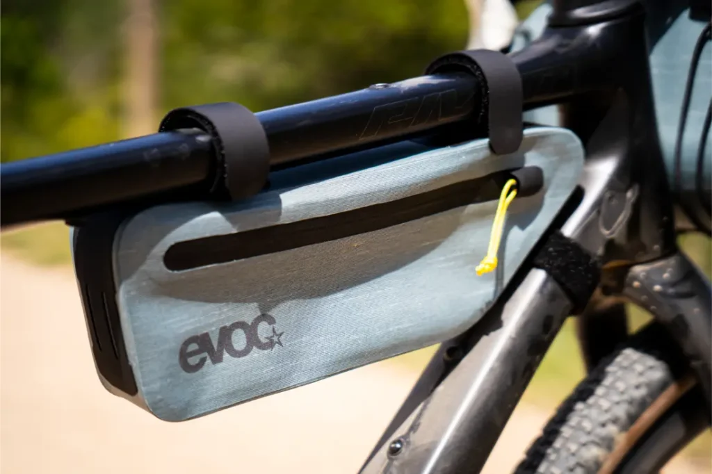 Primer plano de una bolsa para cuadro EVOC de color gris claro, esencial para EVOC Bikepacking, fijada al tubo superior y al tubo inferior de una bicicleta con correas negras de velcro para una mayor protección de la bicicleta.