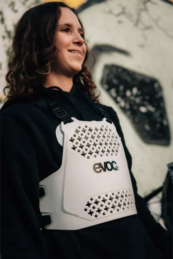 Una persona con una sudadera con capucha negra y una talla XS EVOC Torso Protector blanco se encuentra delante de un mural abstracto, sonriendo.