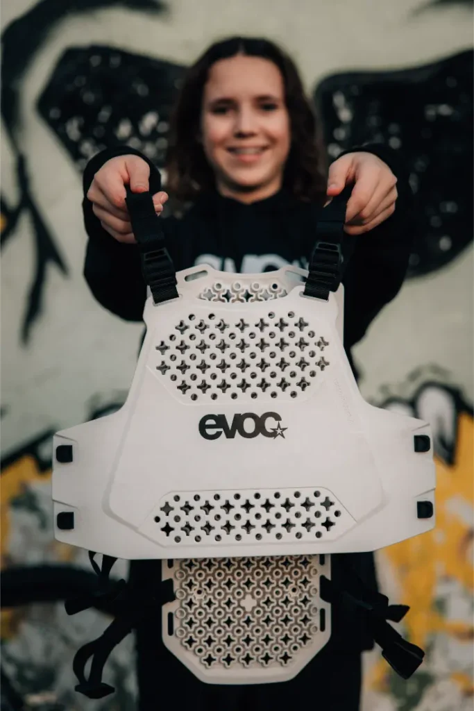 Una persona sostiene un EVOC Torso Protector blanco en talla XS con correas negras, destacando la seguridad, de pie frente a una pared con arte de graffiti negro.
