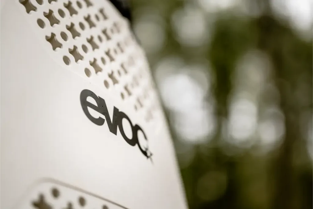 Primer plano de una superficie blanca con orificios de ventilación y el logotipo "evoc" en negro, destacando el EVOC Torso Protector seguridad en talla XS, sobre un fondo verde difuminado.