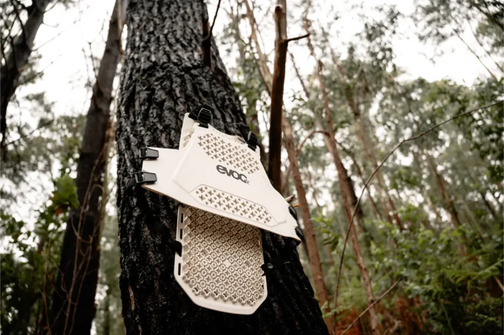Un protector de plástico blanco EVOC Torso Protector, talla XS, con el logotipo "EVOC", se sujeta al tronco de un árbol en una zona boscosa, destacando su enfoque en la seguridad.