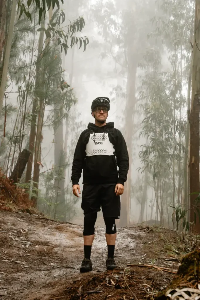 Un hombre vestido con ropa de ciclismo y un protector de torso EVOC (talla XS) se encuentra en un camino de tierra en un bosque neblinoso, rodeado de altos árboles y denso follaje, dando prioridad a la seguridad mientras explora la naturaleza salvaje.