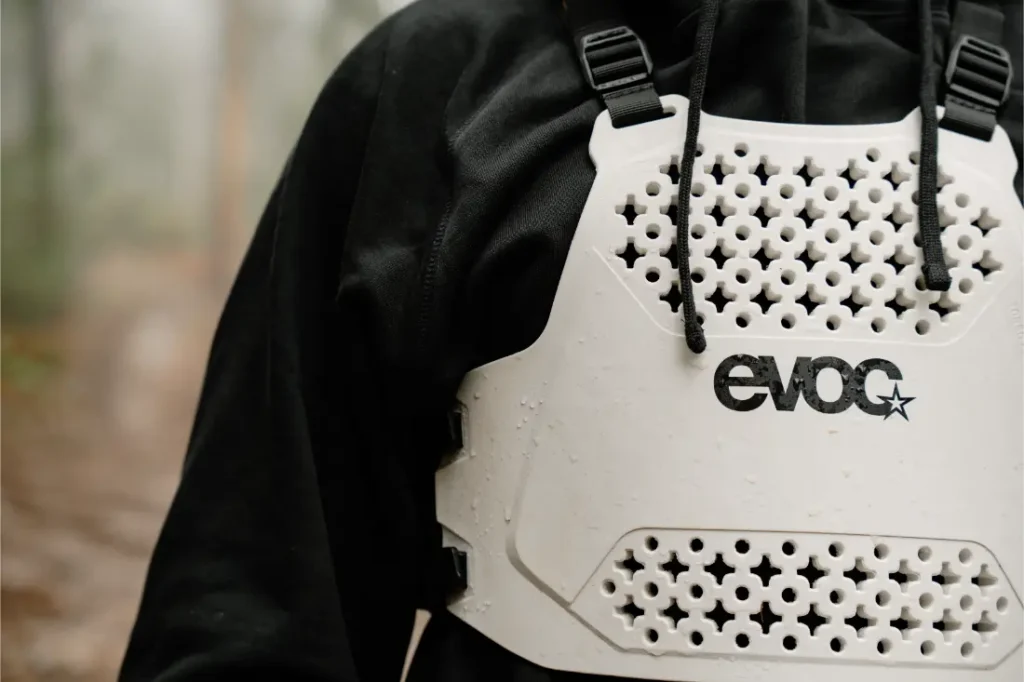 Persona vestida con una sudadera con capucha negra y un EVOC Torso Protector talla XS blanco con un patrón de nido de abeja, de pie al aire libre, mostrando su compromiso con la seguridad.