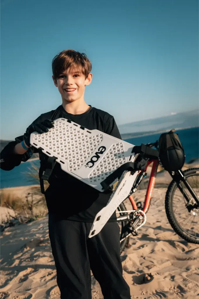 Un niño de pie sobre un terreno arenoso cerca de una playa, sosteniendo una tabla de tracción blanca con la etiqueta "EVOCO", con un EVOC Torso Protector talla XS para seguridad y una bicicleta de montaña al fondo.