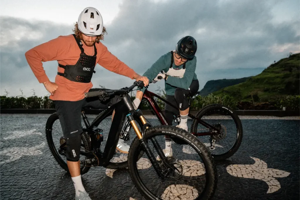 Dos ciclistas con casco, talla XS EVOC Torso Protector y equipo de protección junto a sus bicicletas de montaña en una superficie pavimentada con dibujos de tortugas, al aire libre bajo un cielo nublado, dando prioridad a la seguridad durante su recorrido.