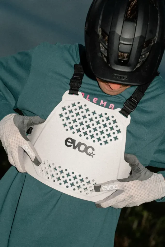 Una persona con casco y guantes ajusta un protector de torso EVOC blanco con diseño perforado, garantizando la seguridad mientras comprueba el ajuste de la talla XS.