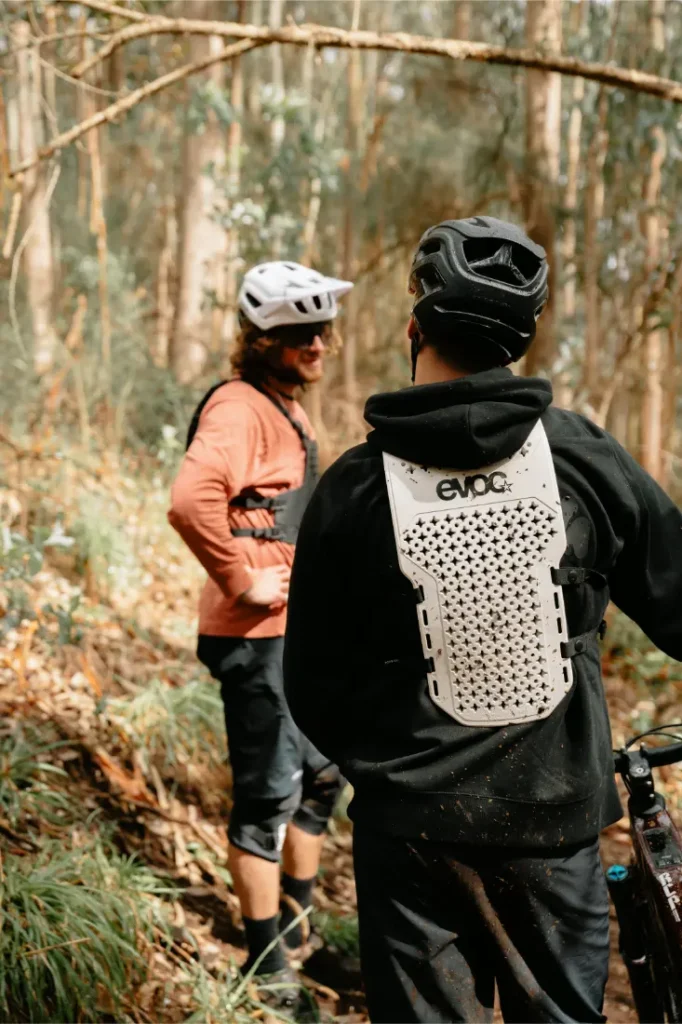 Dos ciclistas con casco y EVOC Torso Protector for seguridad de pie en una pista forestal, uno mirando a la cámara con una bicicleta, el otro girado hacia un lado hablando. Ambos parecen listos para la aventura, con equipo en talla XS.