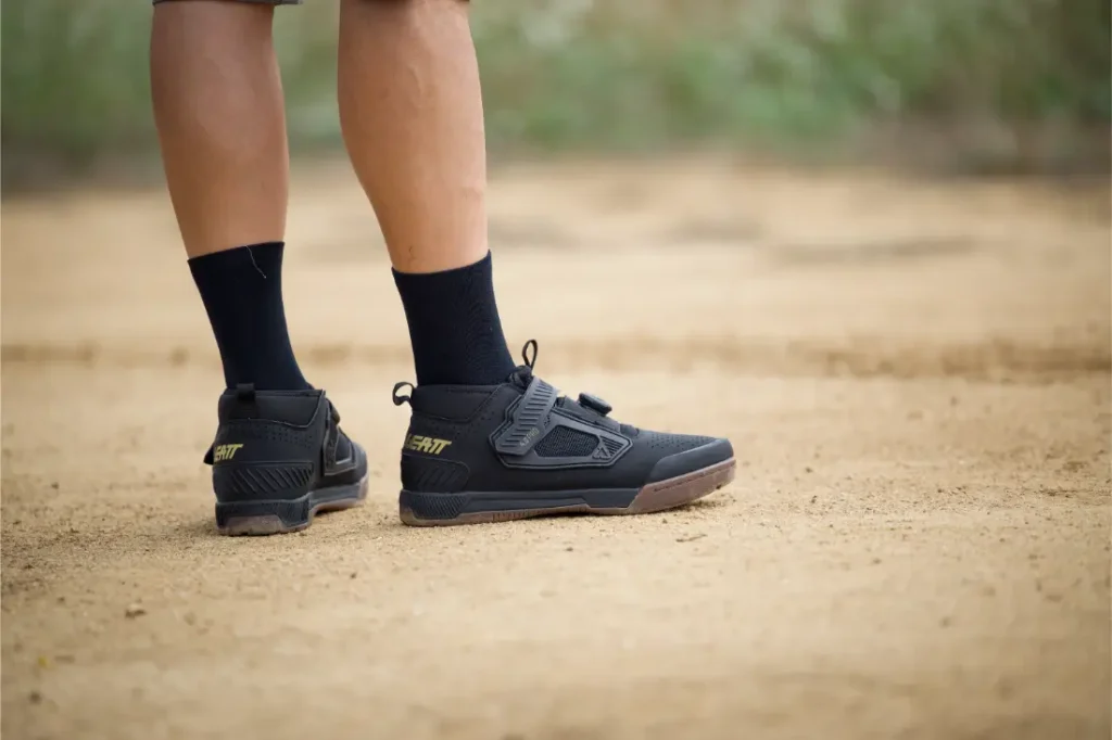 Persona con zapatillas Leatt negras y calcetines de pie en un camino de tierra, en el que sólo se ven las piernas y los pies.