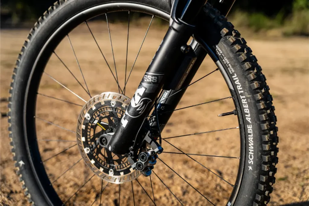 Primer plano de una rueda delantera de bicicleta de montaña con un potente motor Bosch Performance Line CX, freno de disco y cubierta Schwalbe Albert Trail, con un fondo arenoso al aire libre.