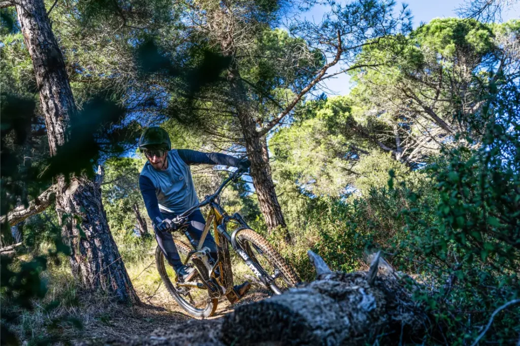 Una persona con casco conduce una bicicleta de montaña equipada con un motor Bosch Performance Line CX por un sendero de tierra a través de una zona boscosa con pinos y la luz del sol filtrándose.
