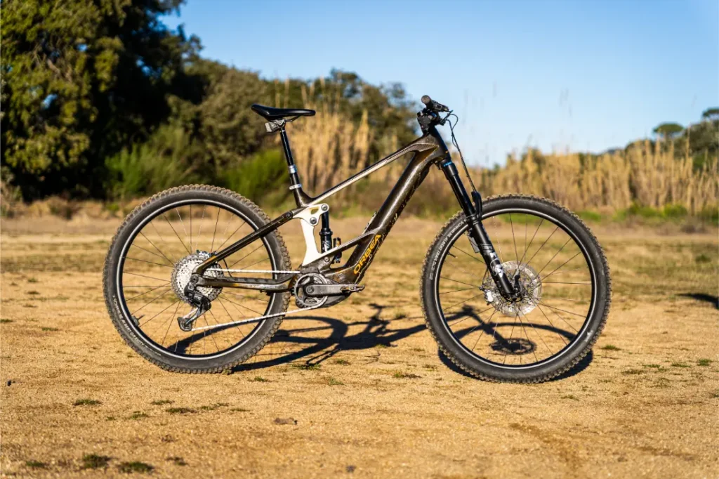 Una bicicleta de montaña con suspensión total y cuadro negro y plateado, equipada con la potente Bosch Performance Line CX, está aparcada en un camino de tierra rodeado de árboles y hierba alta.