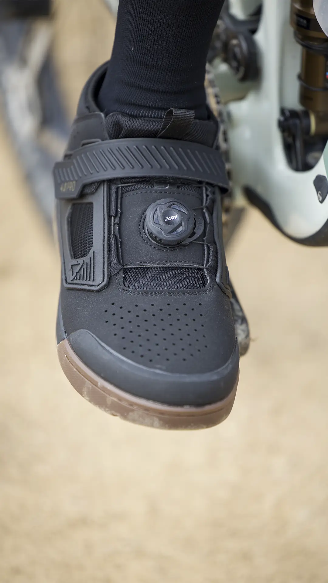 Primer plano de una zapatilla de ciclismo Leatt con sistema de cierre de dial y correa de velcro, llevada por una persona sobre un pedal de bicicleta de montaña.