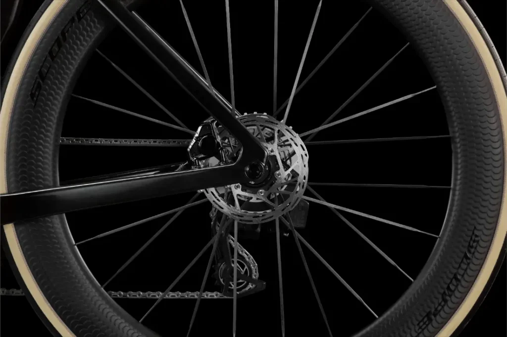Primer plano de una rueda trasera LOOK 795 Blade RS 3 en el que se aprecian el freno de disco, los radios, el cuadro negro, el neumático con flanco claro y parte del mecanismo de cambio, lo que resalta esta bicicleta de carretera de alto rendimiento sobre un fondo negro.