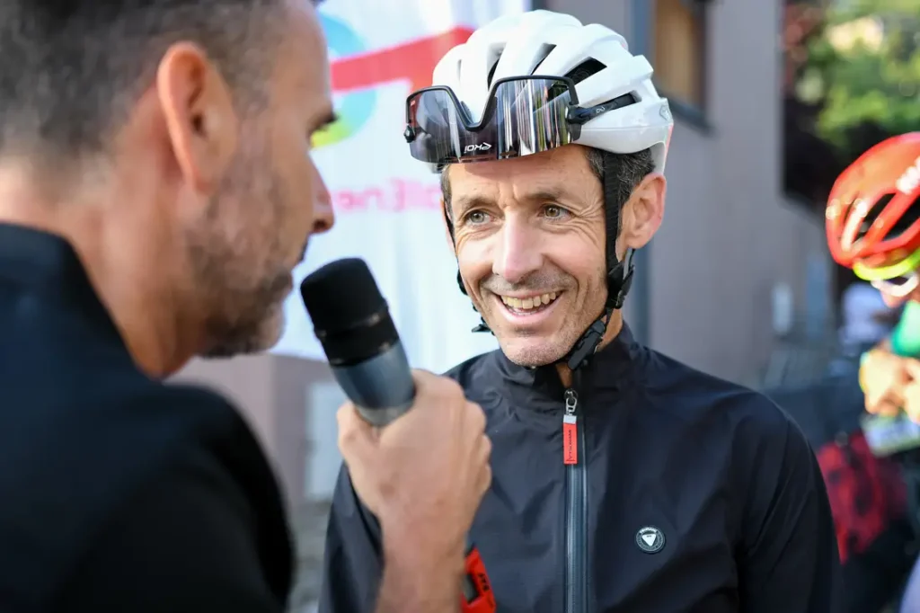 Un hombre con casco ciclista blanco y gafas de sol, posiblemente un cicloturista pirenaico, sonríe mientras es entrevistado por otro hombre que sujeta un micrófono, tal vez después de participar en el evento Alpinum TotalEnergies.