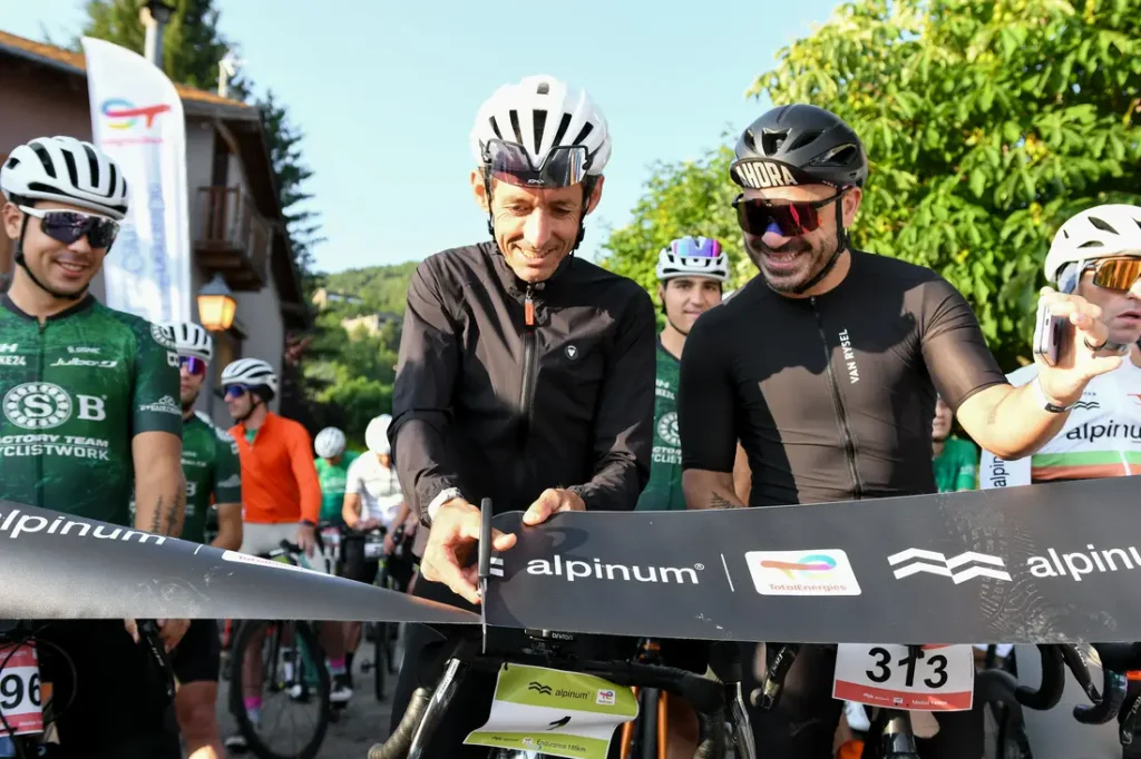 Dos ciclistas con casco y gafas de sol sonríen en la línea de salida de la cicloturista pirenaica, de pie detrás de una barrera con los dorsales y la marca "alpinum", parte de Alpinum TotalEnergies 2026. Otros ciclistas esperan al fondo.