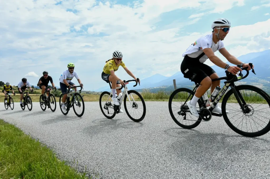 Cinco ciclistas circulan en fila por una carretera rural asfaltada, evocando el espíritu del Tour de Francia, con montañas y un cielo nublado de fondo.