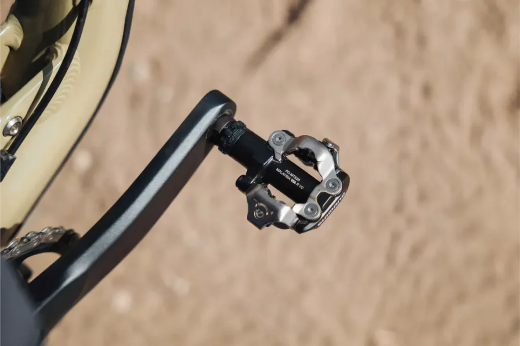 Primer plano de un pedal de bicicleta Shimano DEORE XT con sistema SPD fijado a un brazo de biela, con una superficie de tierra arenosa al fondo, lo que resalta la perfecta configuración de los pedales para senderos.