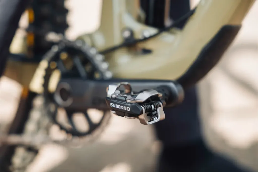 Primer plano de los pedales Shimano DEORE XT Trail acoplados a una biela, con parte del cuadro de la bicicleta y el plato visibles al fondo.