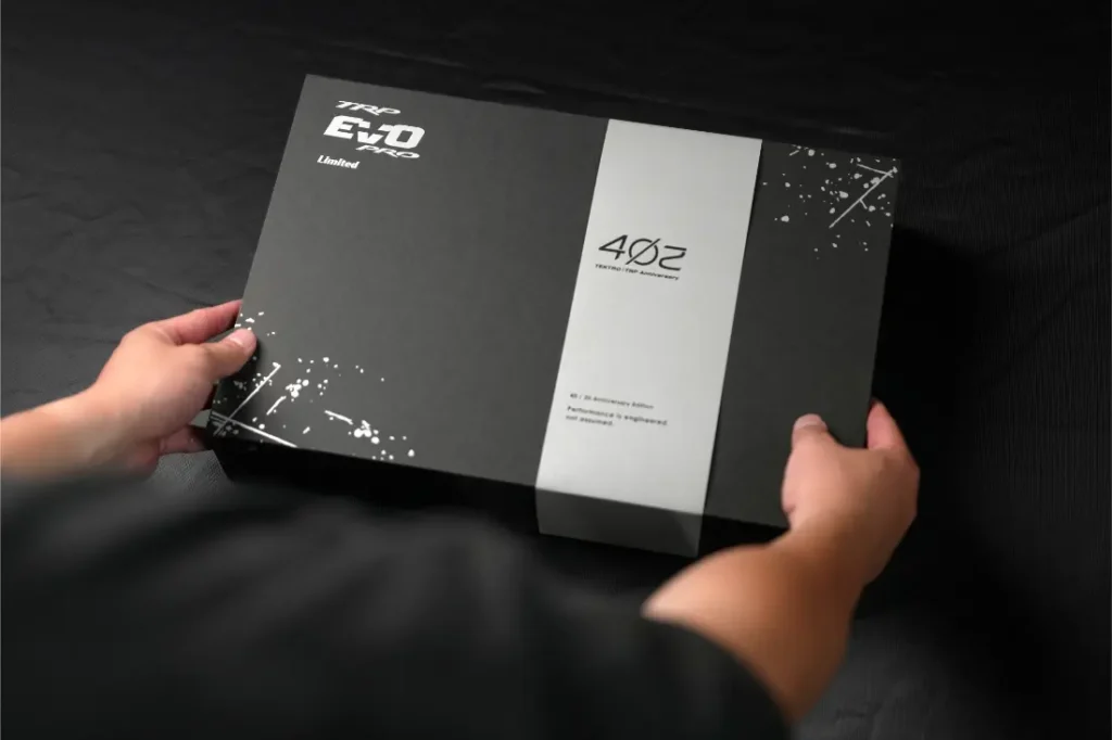 Una persona sostiene una caja rectangular negra con la inscripción «EVO Pro Limited», junto con «4OZ» y elementos decorativos de salpicaduras de pintura blanca: se trata de la TRP EVO PRO Anniversary Edition, una edición limitada exclusiva, expuesta sobre una superficie oscura.