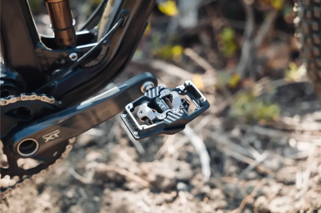 Primer plano de un pedal de bicicleta de montaña, en el que se ven los pedales Shimano DEORE XT SPD Trail acoplados al brazo de la biela, con parte de la cadena y la suspensión trasera visibles en un entorno al aire libre.