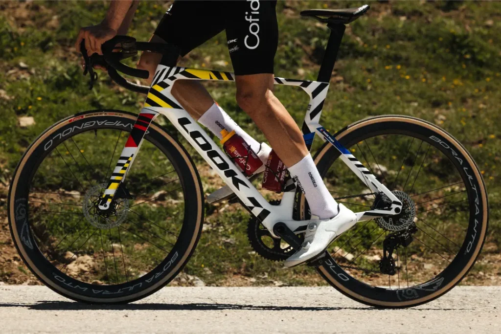 Un ciclista circula en una bicicleta de carretera LOOK 795 Blade RS 3, con un cuadro de diseño colorido, y lleva zapatillas y calcetines blancos, por un camino al aire libre bañado por el sol cerca de Nevers.