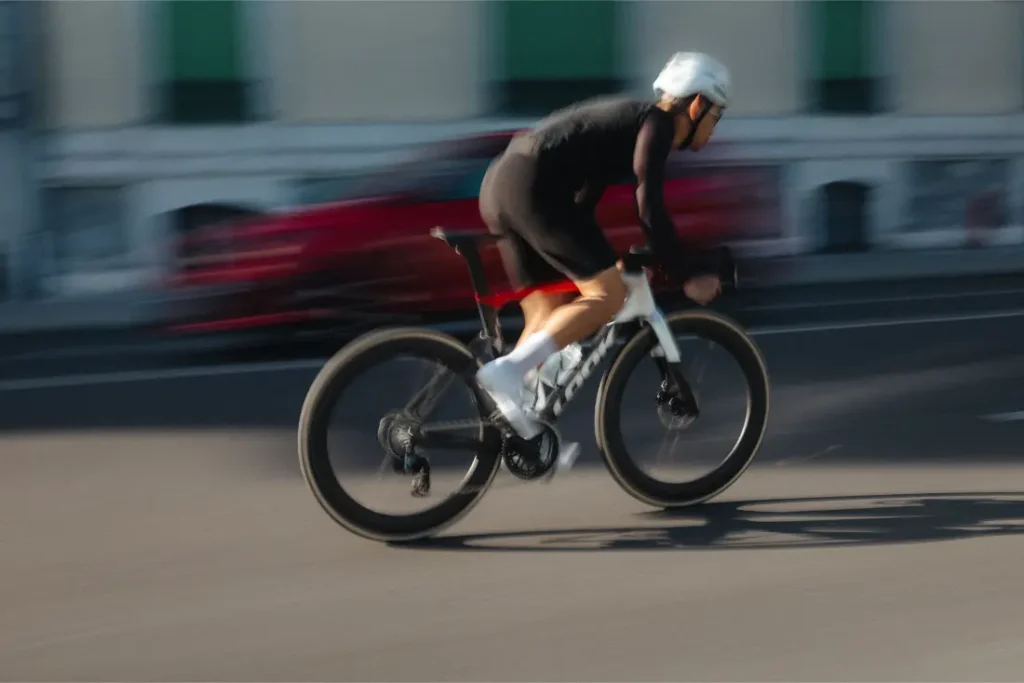 Un ciclista, vestido con ropa negra y casco blanco, recorre a gran velocidad las calles de Nevers montado en una bicicleta de carretera LOOK 795 Blade RS 3, con un coche rojo difuminado al fondo.