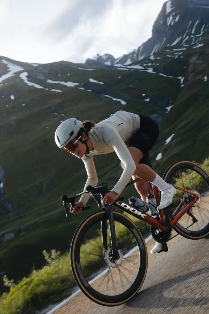 Un ciclista, con casco y gafas de sol, baja en una bicicleta de carretera LOOK 795 Blade RS 3 por una carretera de montaña cerca de Nevers, con picos nevados al fondo.